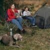 Mor og datter sidder i campingstole med hund, omgivet af natur.