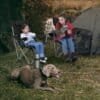 To personer sidder i foldbare campingstole med hund foran telt.