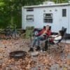 To børn sidder i foldbare campingstole i skov med campingvogn i baggrunden.