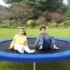 To børn leger på en trampolin i en have med grønne træer og buske.